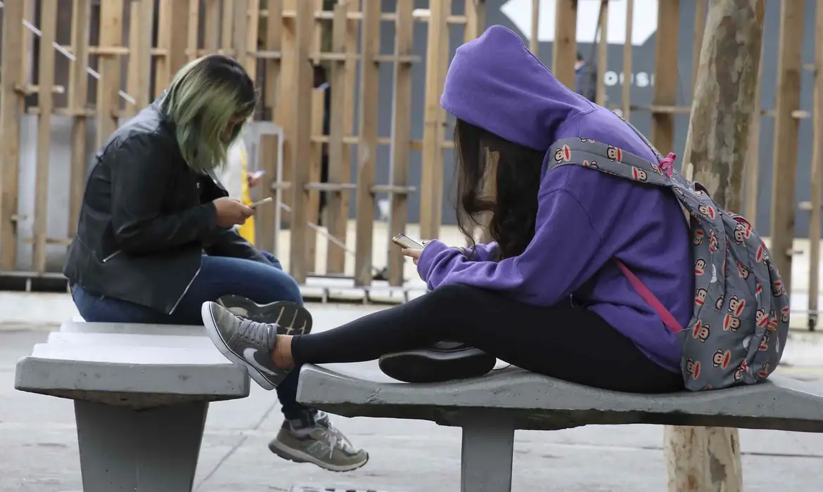 Adolescentes de perfil, sentadas em banco, mexendo no celular