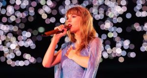 Taylor Swift de perfil, cantando em microfone, com a mão no peito, em show, de cabelo solto e roupa clara