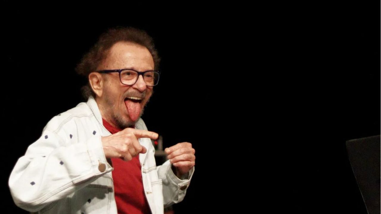 O cantor e compositor Tom Zé fazendo careta e gesticulando sem olhar para a câmera, de óculos