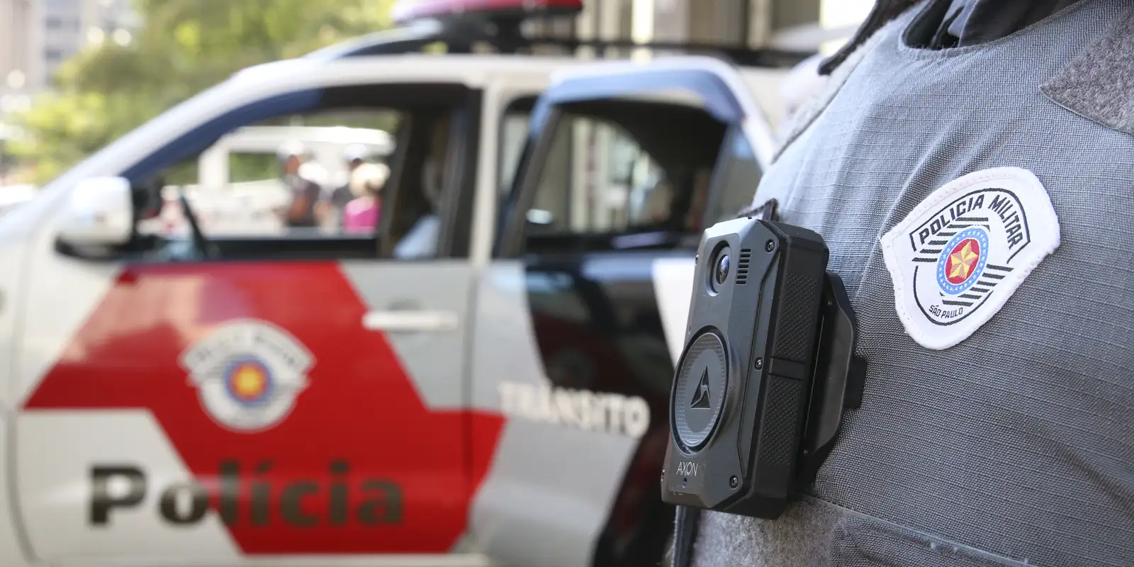Carro da PM de SP ao fundo e agente em primeiro plano sem mostrar o rosto