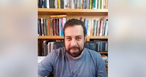 Guilherme Boulos (PSOL) de roupa cinza falando para a câmera com livros ao fundo