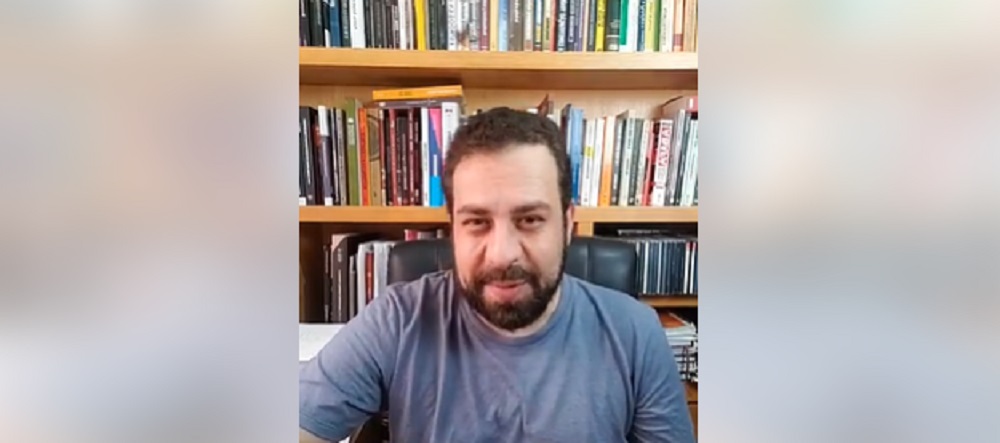 Guilherme Boulos (PSOL) de roupa cinza falando para a câmera com livros ao fundo