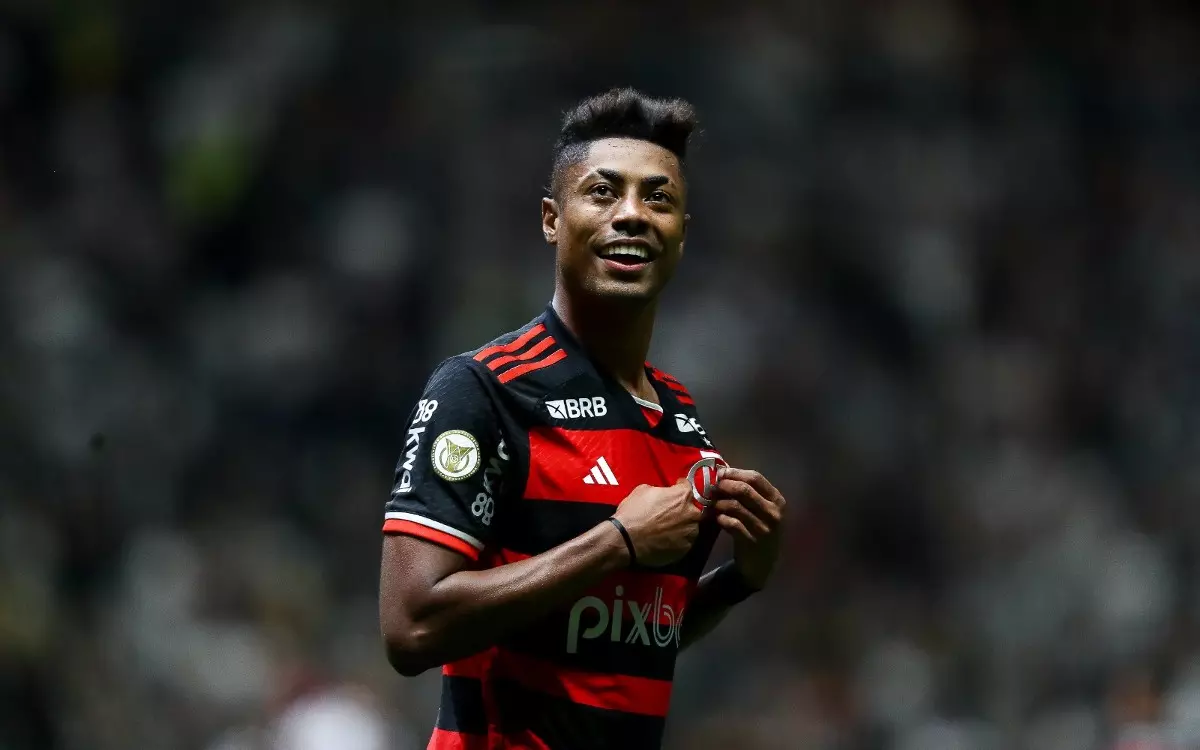 Bruno Henrique, atacante do Flamengo, segurando na camisa do time e sorrindo, em campo