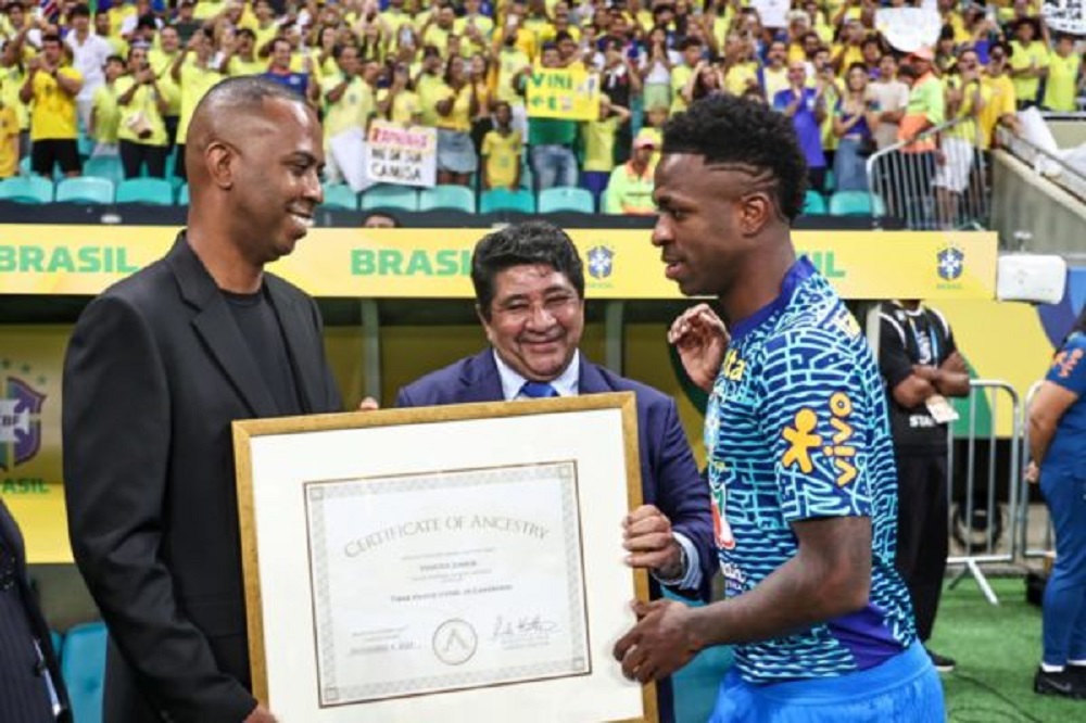 Vinícius Júnior recebendo teste de ancestralidade em estádio lotado, com camisa azul da CBF