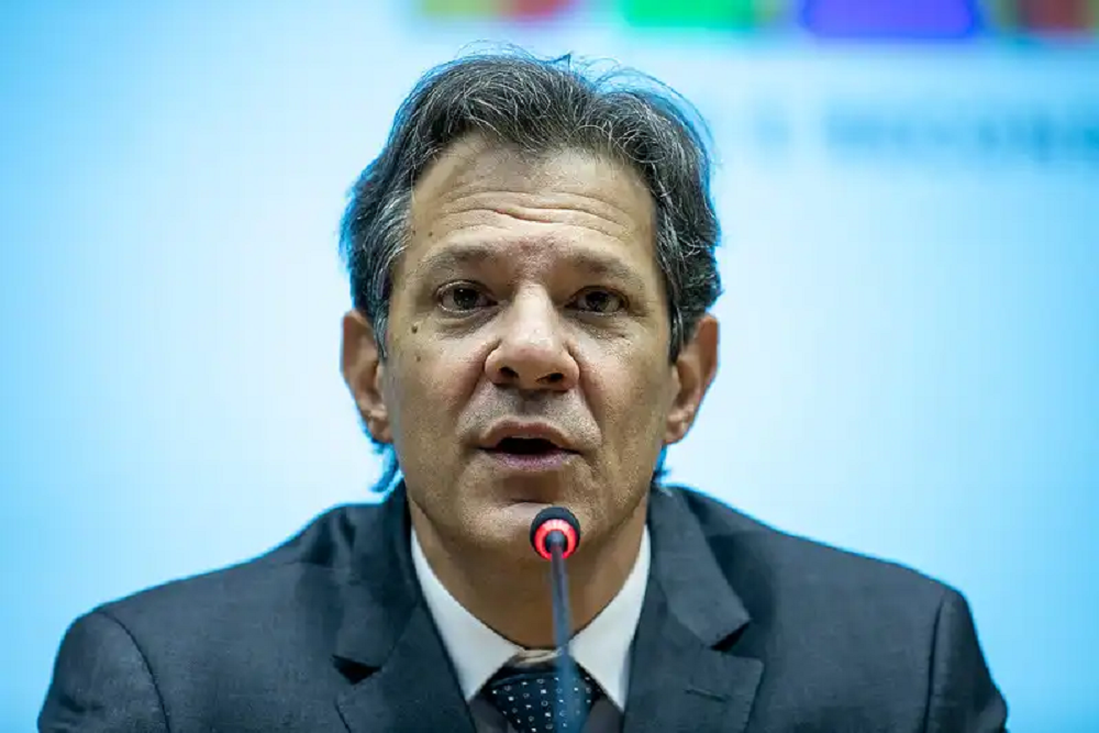 O ministro da Fazenda, Fernando Haddad, olhando pra frente com expressão espantada, perto de microfone