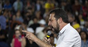 Guilherme Boulos de lado, falando em microfone e apontando