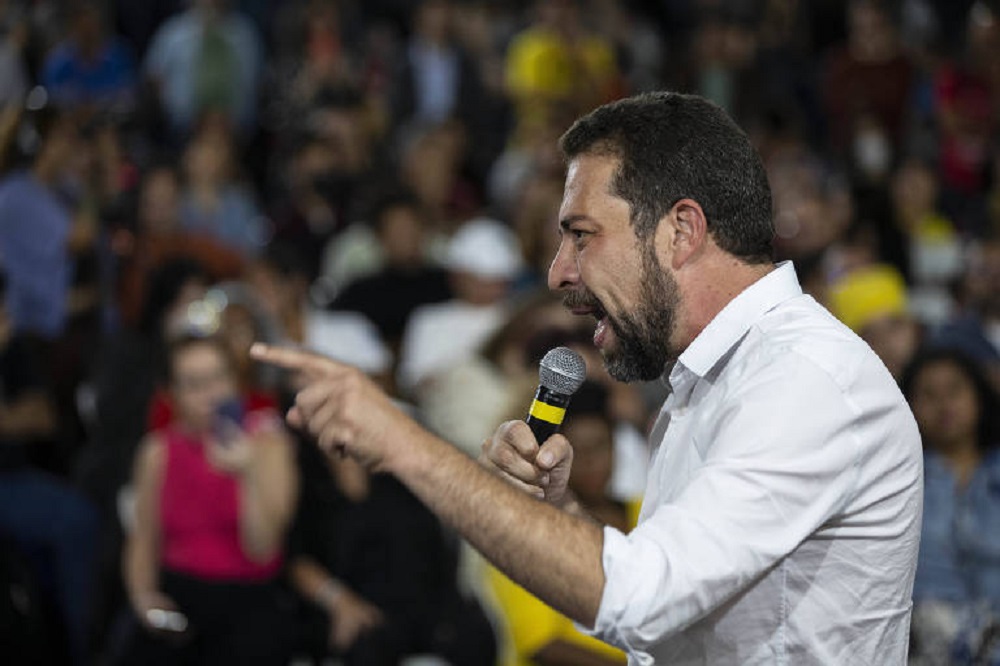 Guilherme Boulos de lado, falando em microfone e apontando