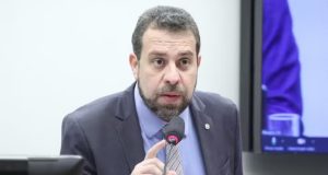 deputado federal Guilherme Boulos (PSOL-SP) falando em microfone com expressão assustada sem olhar para a câmera, de terno e gravata em tons de azul e bege
