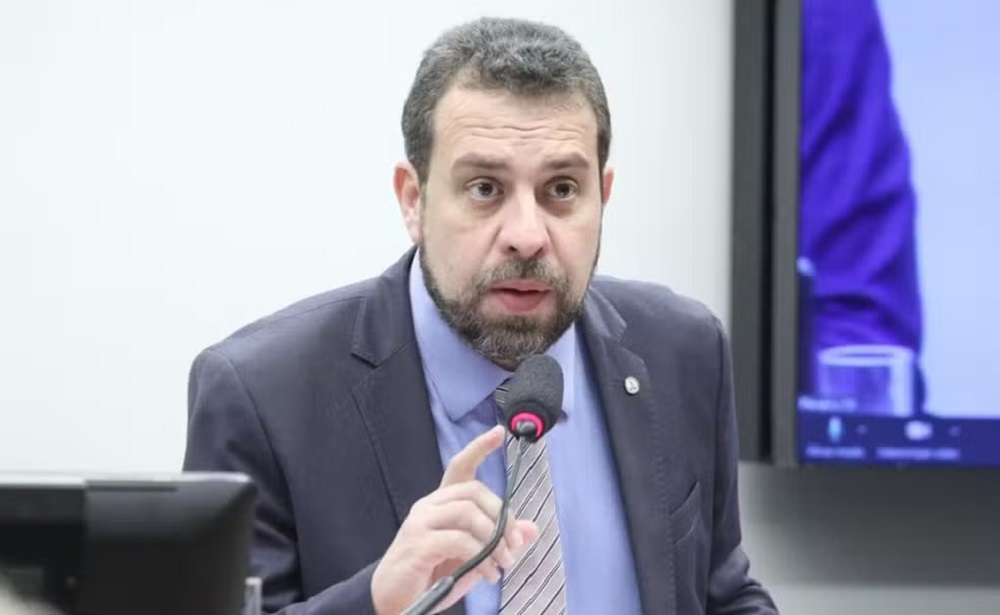 deputado federal Guilherme Boulos (PSOL-SP) falando em microfone com expressão assustada sem olhar para a câmera, de terno e gravata em tons de azul e bege