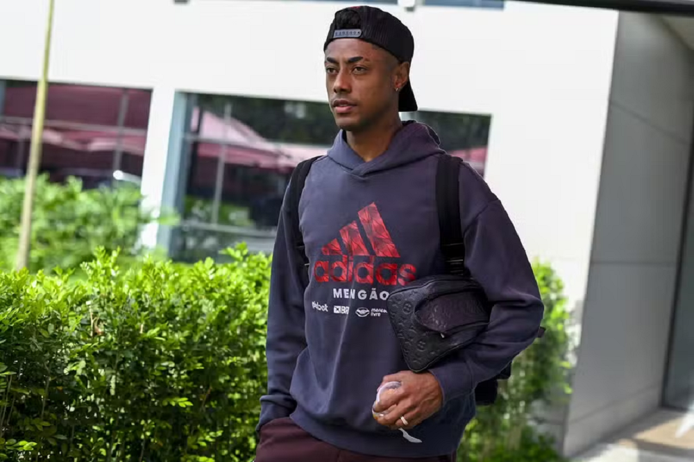 Bruno Henrique com moletom cinza do Flamengo, boné preto virado para trás e mochila preta nas costas, caminhando, sem olhar para a câmera