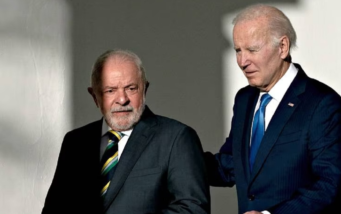 Joe Biden caminhando atrás de Lula, com as mãos nas costas dele, ambos sérios