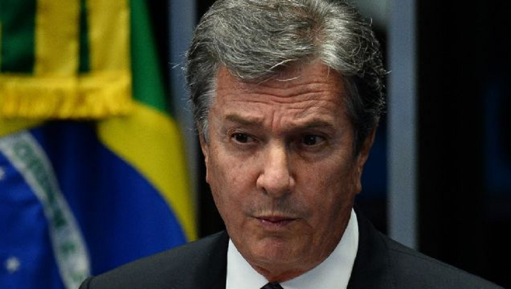 Fernando Collor de Mello com expressão pensativa, em close, com bandeira do Brasil ao fundo
