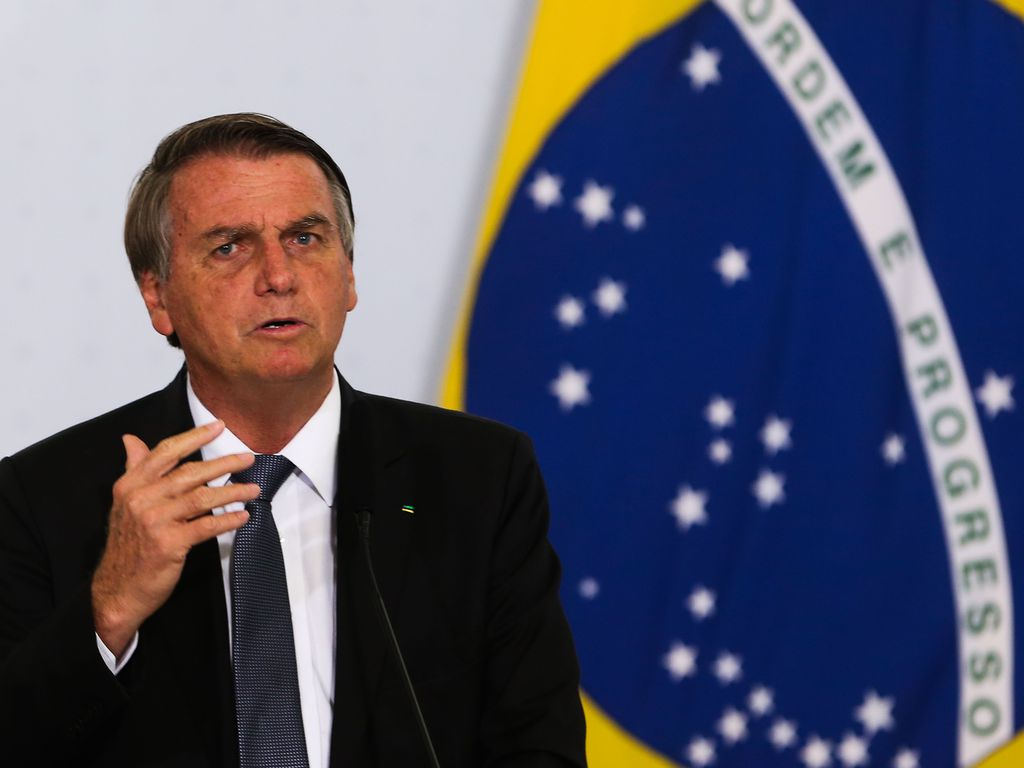 Jair Bolsonaro falando e gesticulando, sério, com bandeira do Brasil ao fundo