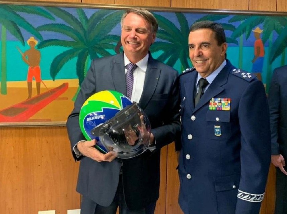 Jair Bolsonaro segurando capacete ao lado de Carlos Baptista Jr., ambos em pé e sorrindo sem olhar para a câmera