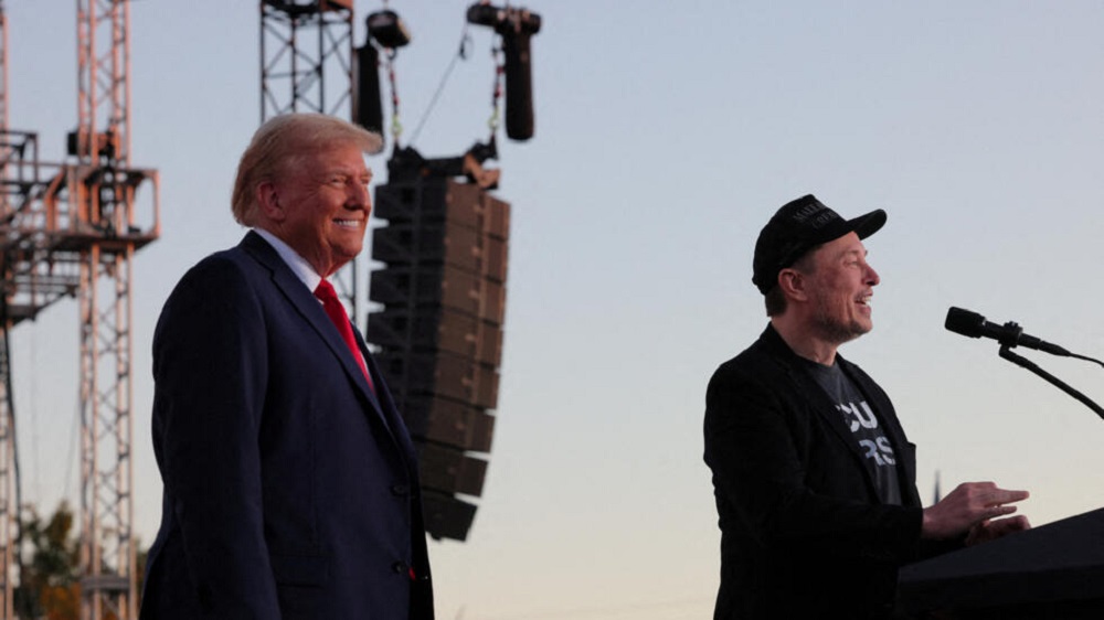 Donald Trump e Elon Musk em palco, o primeiro sorrindo e o segundo falando em microfone