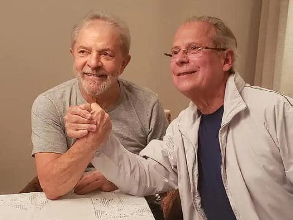 Lula e José Dirceu sorrindo em aperto de mãos, ambos sem olhar para a câmera