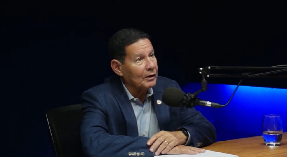 O senador Hamilton Mourão (Republicanos-RS) falando em microfone em estúdio de podcast, com roupa social azul e preta, sem olhar para a câmera, sério e sentado