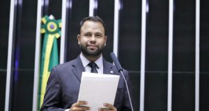 deputado Pastor Henrique Vieira (RJ) sorrindo na Câmara, segurando papel, sem olhar para a câmera, em pé