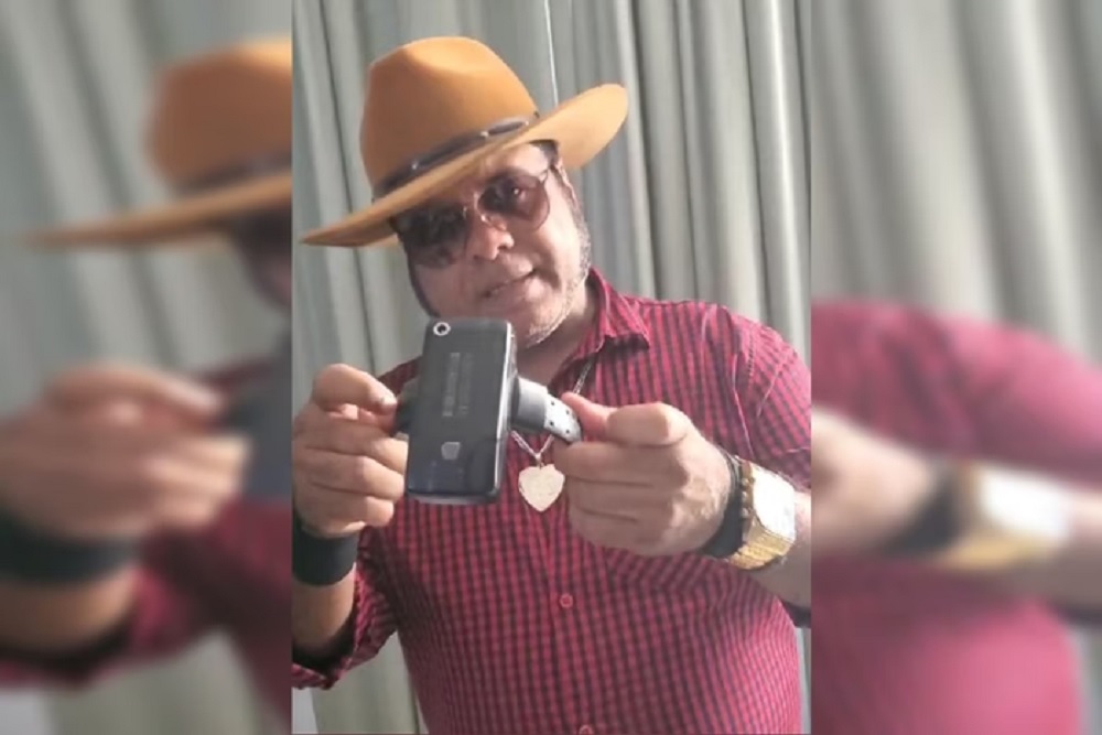 Roque Saldanha no centro de montagem, segurando tornozeleira eletrõnica, de chapéu e óculos escuros