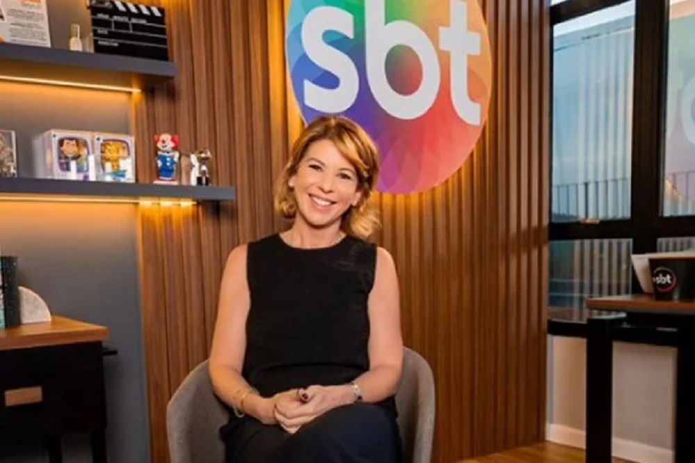 Daniela Beyruti de roupa preta posando sentada perto de símbolo do SBT