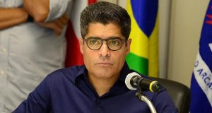 ex-prefeito de Salvador ACM Neto de óculos de grau e camisa social azul, sério, com bandeiras e pessoas ao fundo