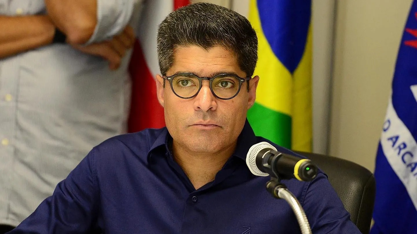 ex-prefeito de Salvador ACM Neto de óculos de grau e camisa social azul, sério, com bandeiras e pessoas ao fundo