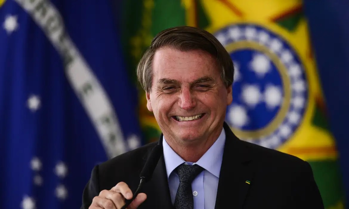 ex-presidente Jair Bolsonaro (PL) rindo e segurando microfone, com bandeiras ao fundo