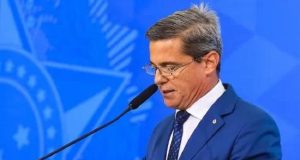 General da reserva Mario Fernandes de perfil e de roupa social azul, falando em microfone, cabisbaixo e sério