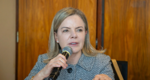 Gleisi Hoffmann de roupa social, falando em microfone, de cabelo solto, séria, gesticulando
