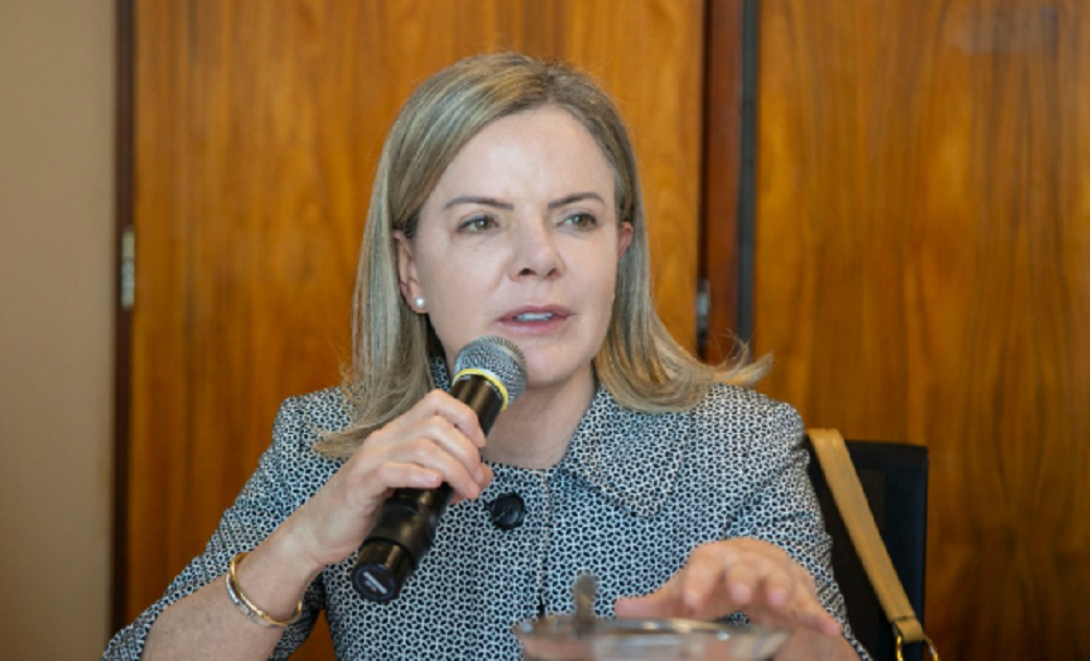 Gleisi Hoffmann de roupa social, falando em microfone, de cabelo solto, séria, gesticulando