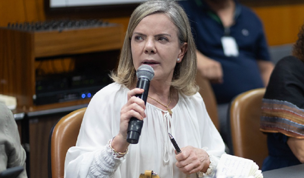 A deputada federal Gleisi Hoffmann (PT) de cabelo solto e roupa branca, falando em microfone, séria, olhando para o lado, sentada