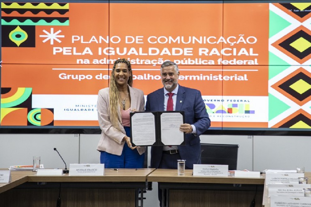 A ministra da Igualdade Racial, Anielle Franco, e o ministro da Secom, Paulo Pimenta, sorrindo lado a lado para foto exibindo documentos