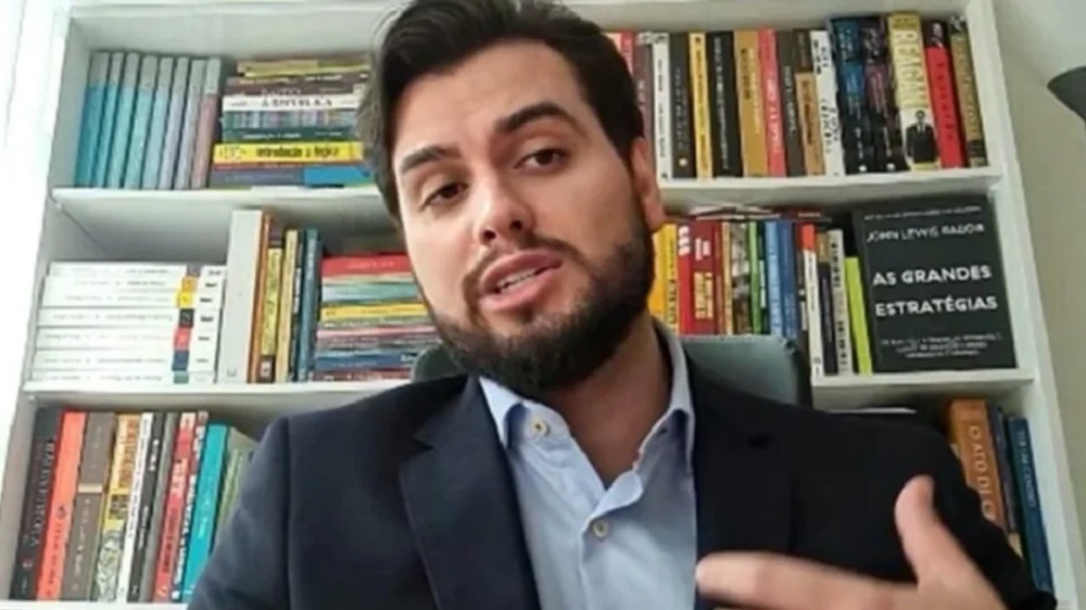 Filipe Garcia Martins na frente de estante cheia de livros, falando e gesticulando, de roupa social, sem olhar para a câmera