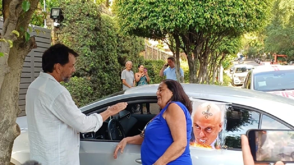 Idosa de roupa azul conversando com homem de branco perto de carro