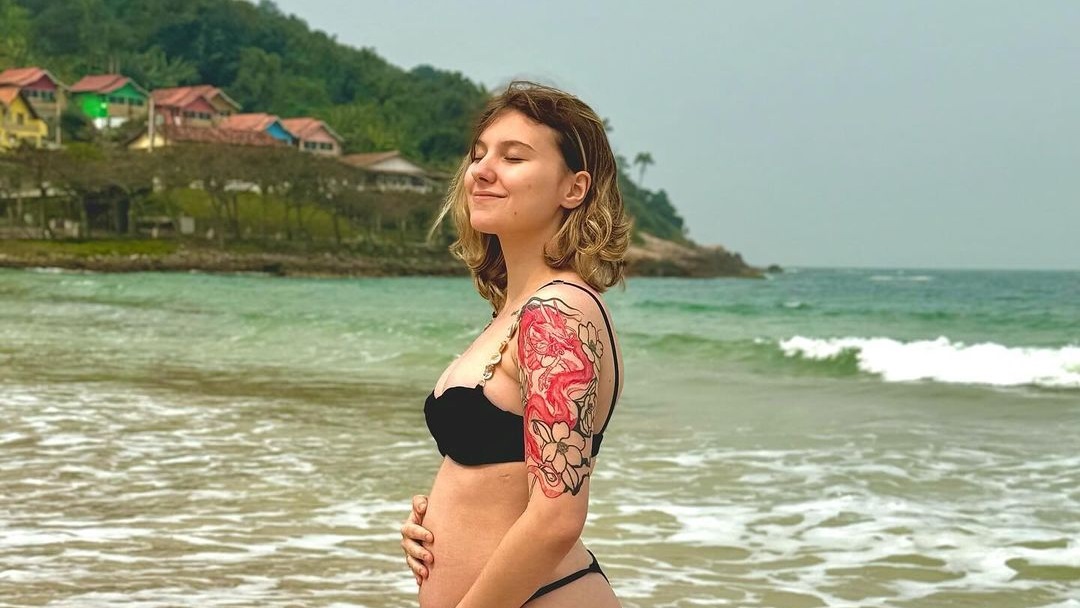 Isabel Veloso de biquíni preto, de perfil, com mão na barriga grávida, na praia, com expressão serena