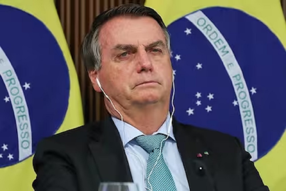 Jair Bolsonaro (PL) com cara de choro, usando fones de ouvido brancos, com bandeiras ao fundo