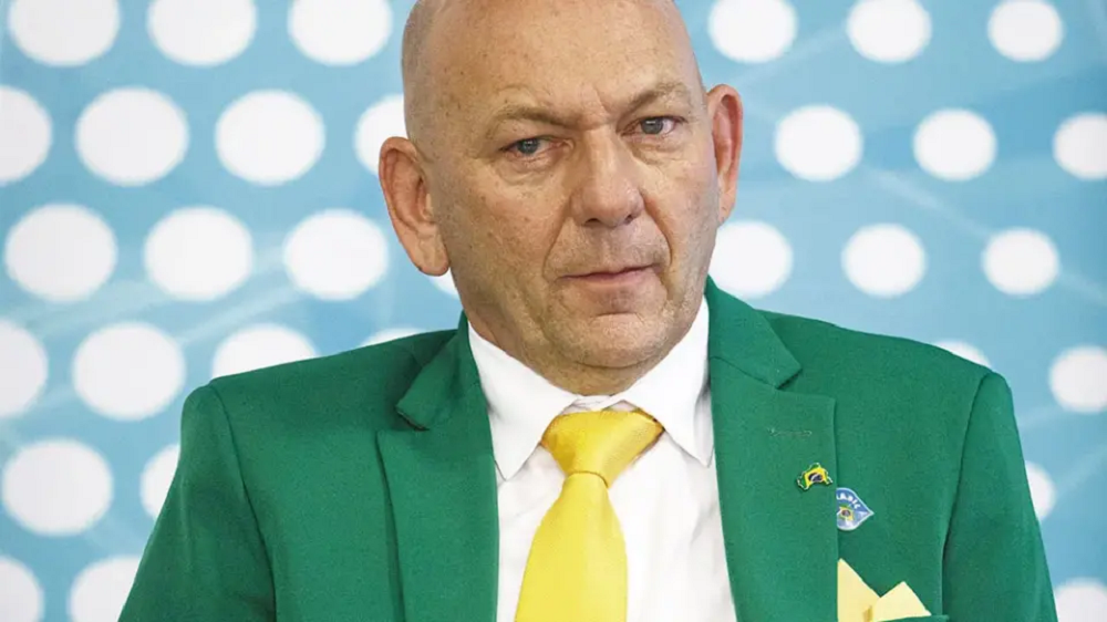 Luciano Hang de roupa verde e amarela, sério, em fundo de foto azul com bolinhas brancas
