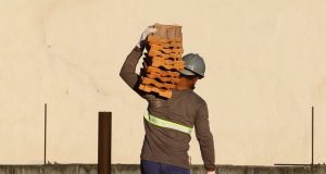 Homem de costas em construção, de capacete de proteção, segurando tijolos