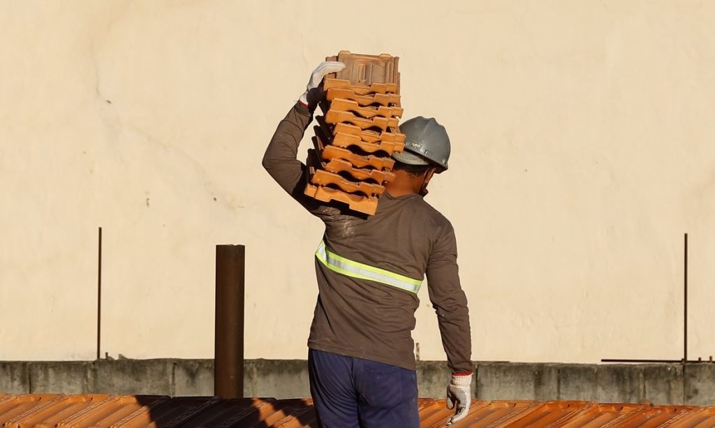 Homem de costas em construção, de capacete de proteção, segurando tijolos