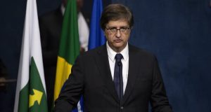 O procurador-geral da República, Paulo Gonet, falando em microfone, sério, de óculos, com bandeiras ao fundo