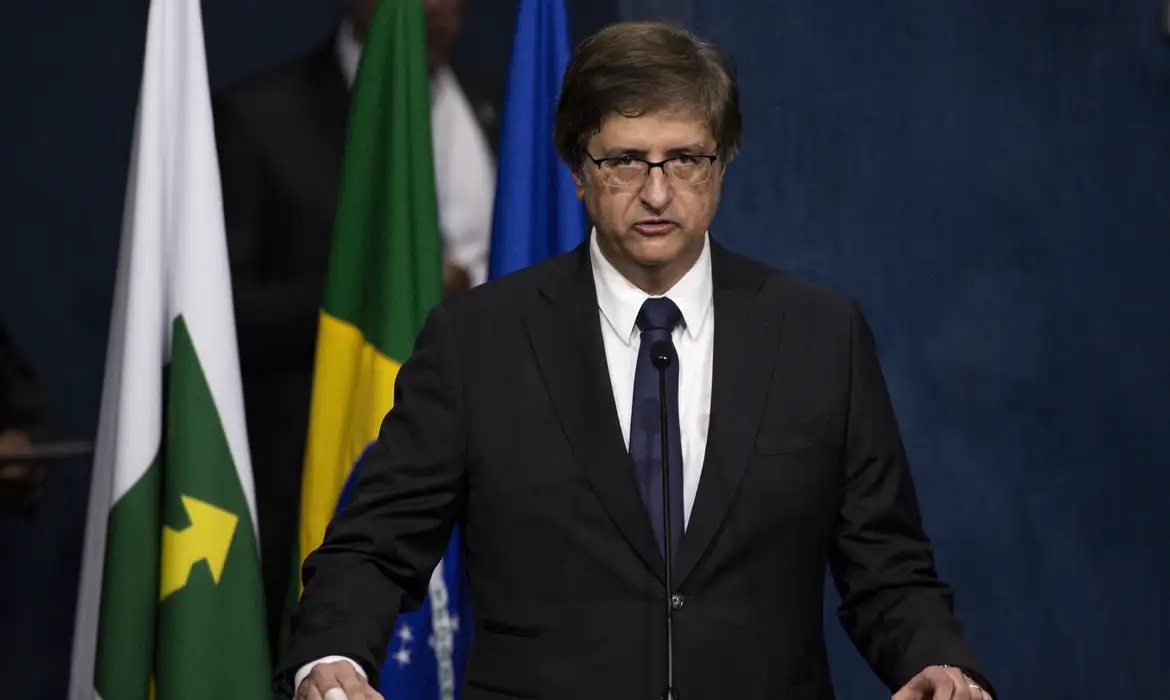 O procurador-geral da República, Paulo Gonet, falando em microfone, sério, de óculos, com bandeiras ao fundo