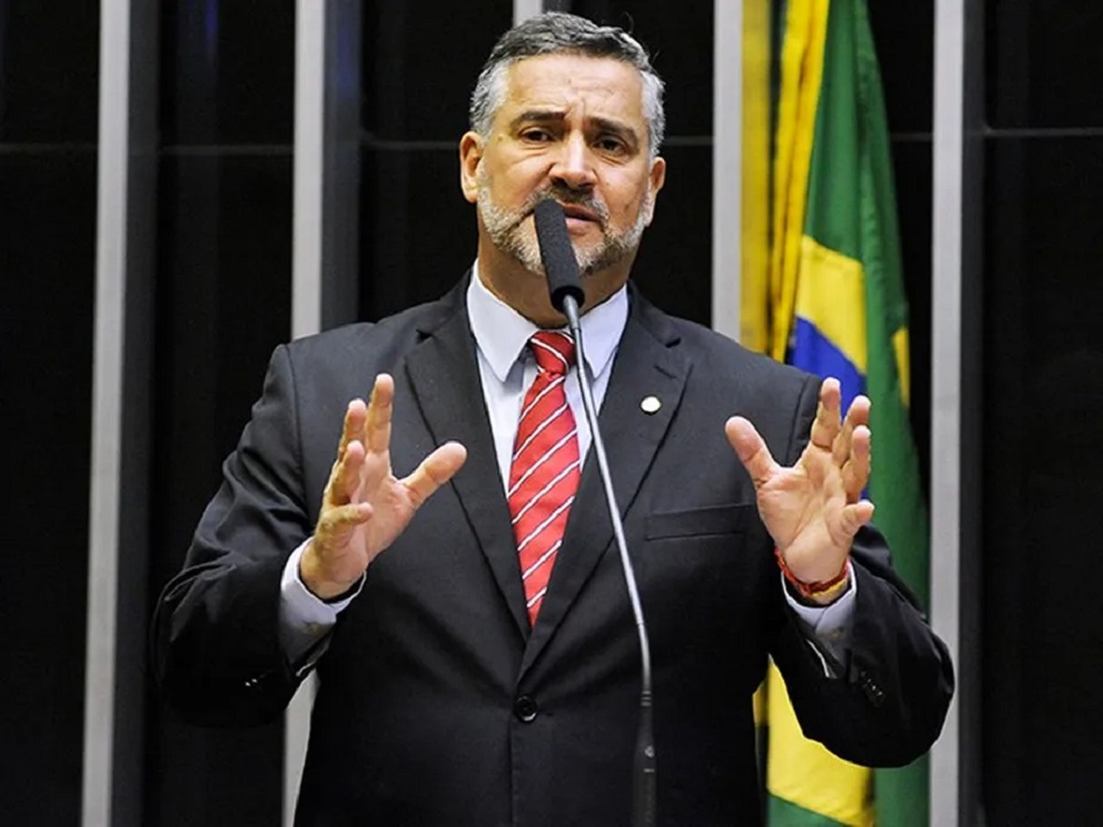 Paulo Pimenta, Ministro-Chefe da Secom do Governo Lula, falando e gesticulando na Câmara, sério, com bandeiras ao fundo
