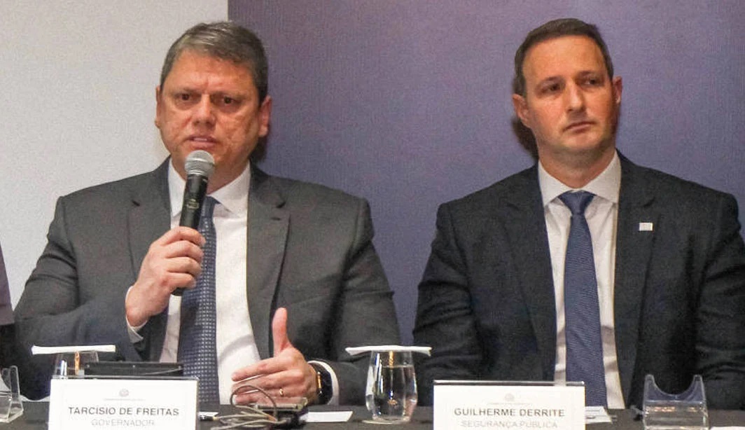 O governador Tarcísio de Freitas (Republicanos) e o secretário da Segurança Pública Guilherme Derrite lado a lado, sentados, o primeiro falando e o segundo sério
