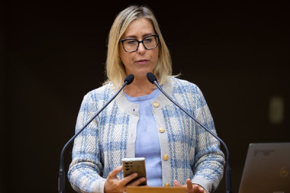 Vereadora Fernanda Barth (PL) de roupa azul e óculos de grau com armação preta, de cabelo solto, falando em microfone e segurando celular, séria