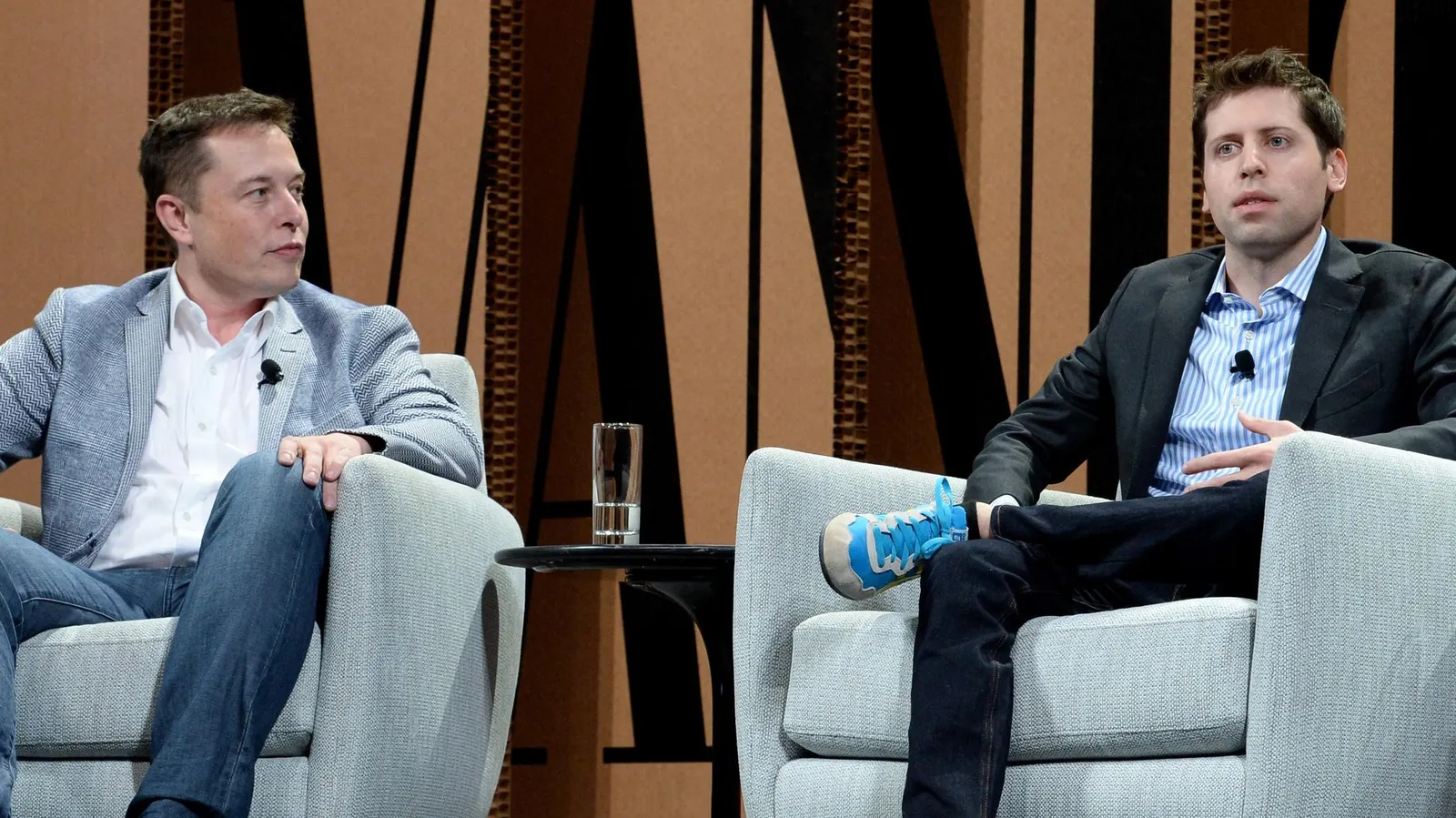 Elon Musk e Sam Altman sentados lado a lado, sérios, falando