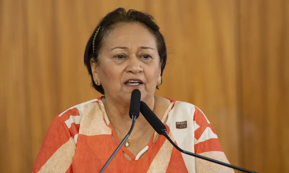 A governadora do Rio Grande do Norte, Fátima Bezerra (PT), falando em microfones, séria, de roupa estampada