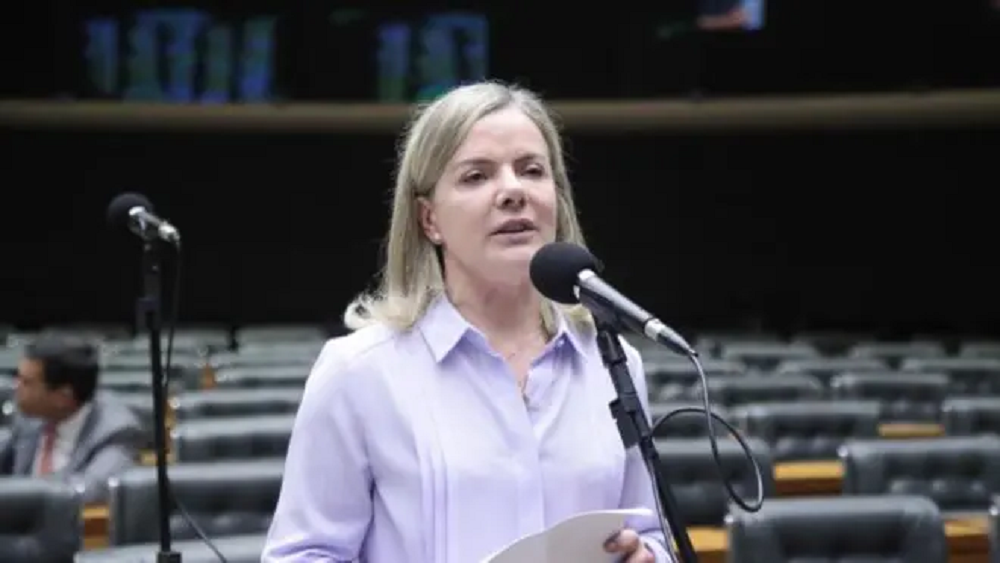 A presidente do PT, Gleisi Hoffmann, de roupa social azul clara e cabelo solto, segurando papel e falando em microfone na Câmara dos Deputados