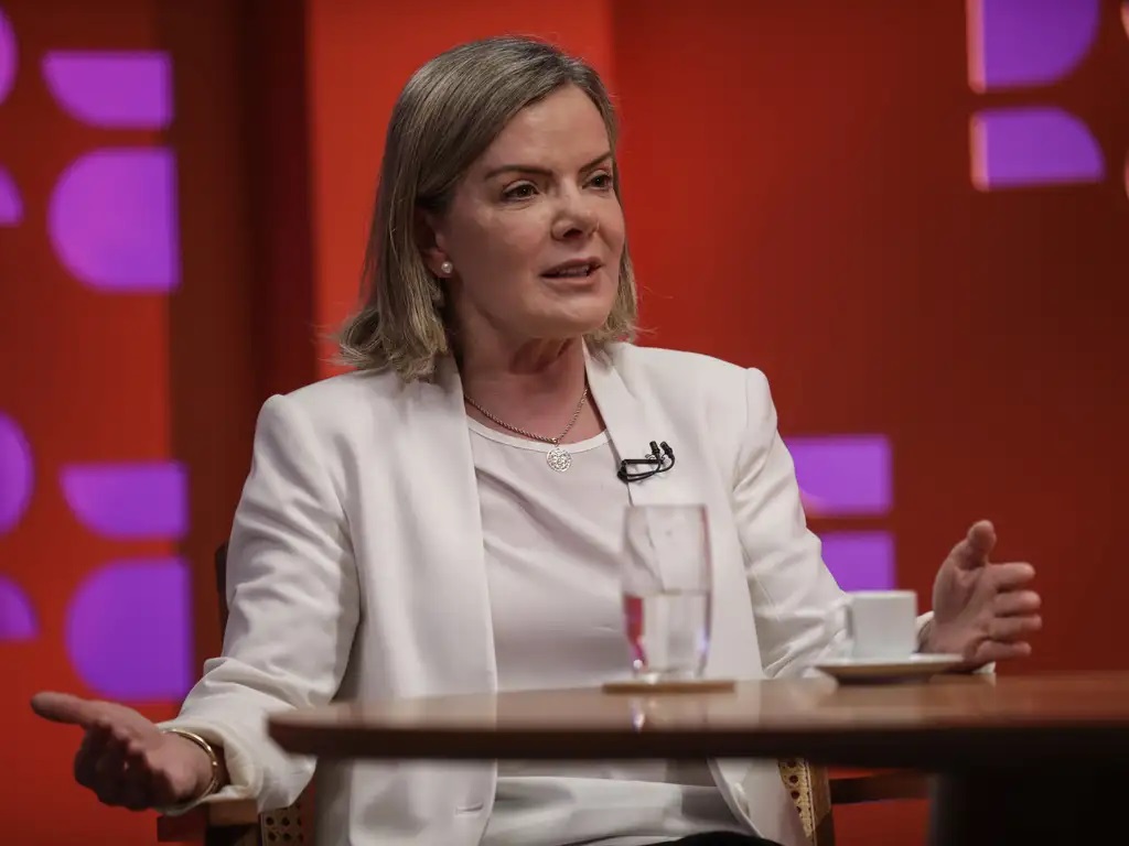 A presidente do PT, Gleisi Hoffmann, de roupa social clara, falando e gesticulando, séria, sentada perto de mesa, de cabelo solto
