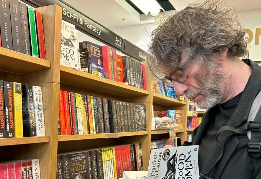 Neil Gaiman em biblioteca, cabisbaixo, lendo livro