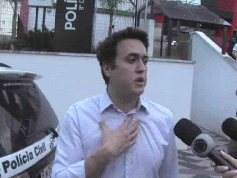 Vinícius Gritzbach de camisa social branca, com a mão no peito, falando, sério, em entrevista, perto de carro da polícia